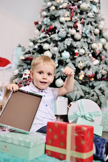 Le petit garçon ouvre des boîtes avec des cadeaux sous le sapin de Noël et sourit