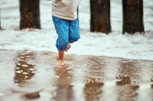 Petit garçon en pantalon bleu courant sur la côte de la mer et jouant avec les vagues de la mer jambes floues floues Petit garçon courant sur la plage à travers des éclaboussures et des gouttes d'eau de mer