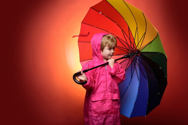 Petit garçon avec parapluie de couleur arc-en-ciel isolé sur fond rouge. Enfant annoncez vos produits et services. Mignon petit garçon enfant portant des vêtements d'automne le jour de la pluie d'automne. Enfant sous la pluie.