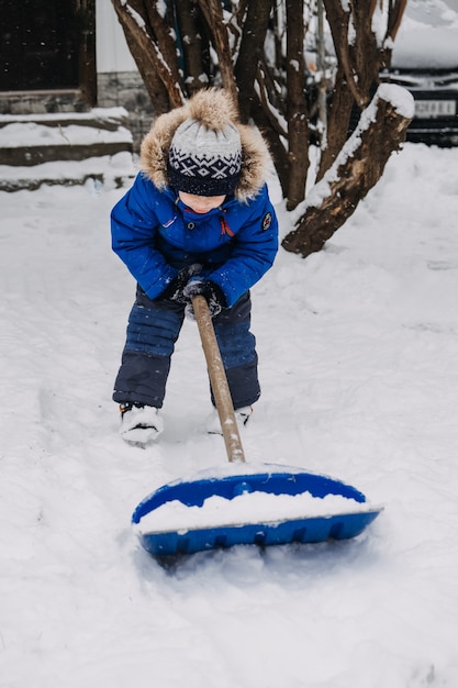 Petit garçon avec une pelle à la main enlever la neige dans l'arrière-cour de déneigement kid en veste bleue nettoie la neige