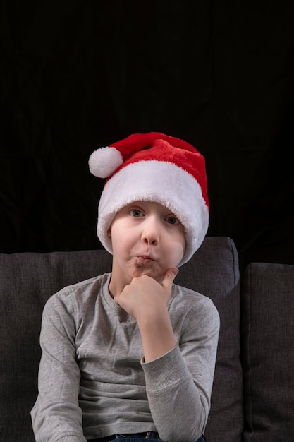 Petit garçon pensif en bonnet de Noel assis sur le canapé sur fond noir.