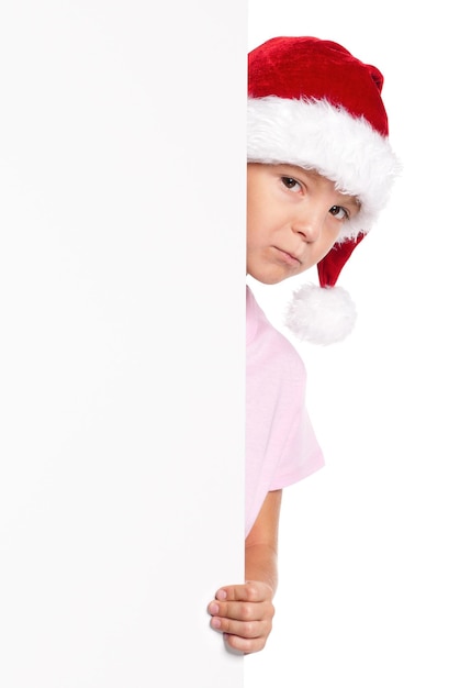 Petit garçon portant un chapeau de Père Noël posant derrière un tableau blanc isolé sur un fond blanc Enfant mignon tenant un panneau d'affichage de Noël vide Enfant regardant derrière un panneaux de vacances vides