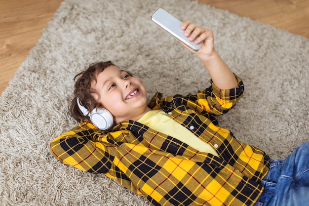 Petit garçon positif portant des écouteurs faisant selfie sur smartphone souriant à l'appareil allongé sur l'espace libre du sol