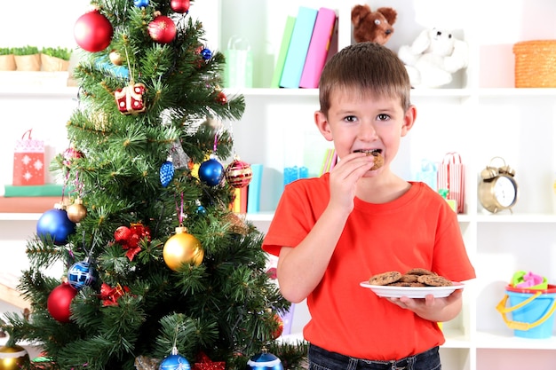 Le petit garçon près de l'arbre de Noël mange des biscuits