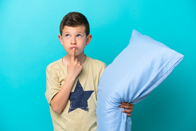 Petit garçon en pyjama isolé sur fond bleu ayant des doutes en levant les yeux