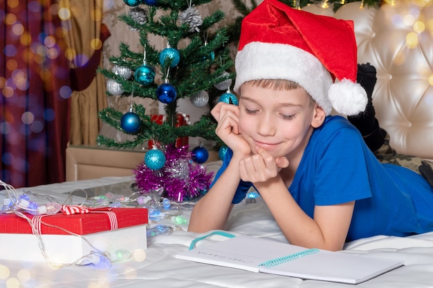 Un petit garçon de race blanche dans un chapeau de Père Noël rouge se trouve à rêver de cadeaux et propose une lettre au Père Noël à la maison décorée pour Noël. Traditions autour du Nouvel An et des vacances de Noël