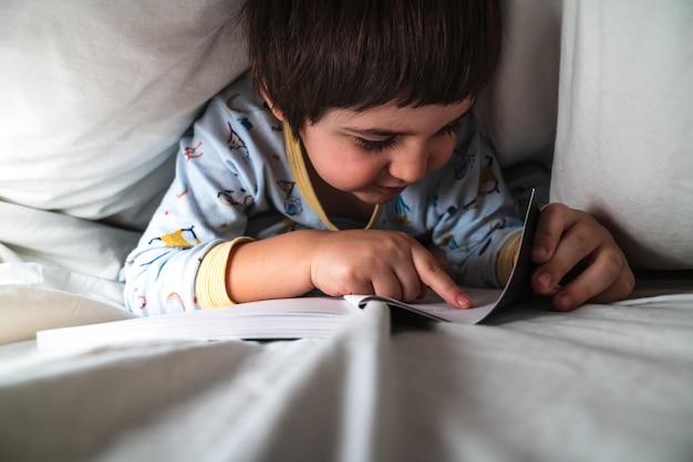 Petit garçon recouvert des draps de son lit la nuit avec son pyjama, lisant joyeusement un livre