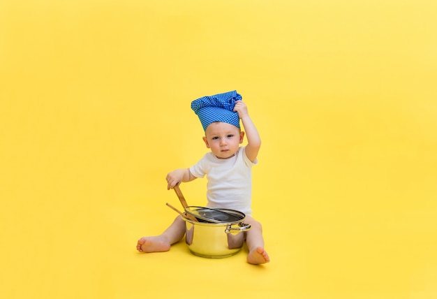 Un petit garçon regarde et une toque avec un pot et une cuillère à spatule en bois. Un garçon dans un body blanc tire sur une casquette