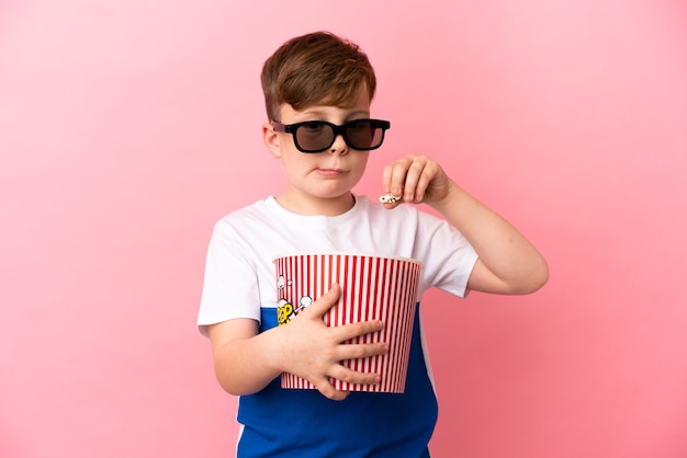 Petit garçon roux isolé sur fond rose avec des lunettes 3d et tenant un grand seau de pop-corn