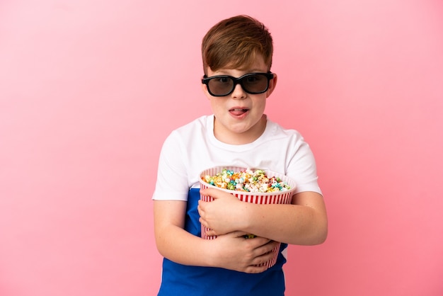 Petit garçon roux isolé sur fond rose avec des lunettes 3d et tenant un grand seau de pop-corn