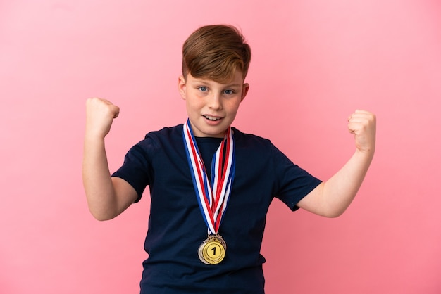 Petit garçon roux avec des médailles isolées sur fond rose célébrant une victoire