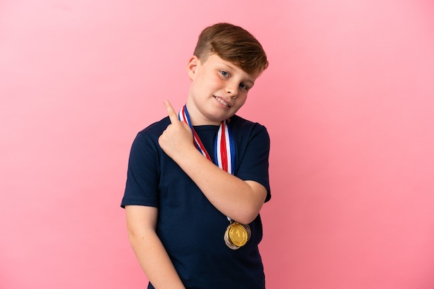 Petit garçon roux avec des médailles isolées sur fond rose pointant sur le côté pour présenter un produit