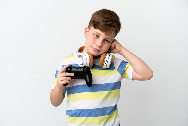 Petit garçon roux tenant une manette de jeu isolé sur fond blanc frustré et couvrant les oreilles