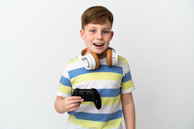 Petit garçon roux tenant une manette de jeu isolée sur un mur blanc avec une expression faciale surprise