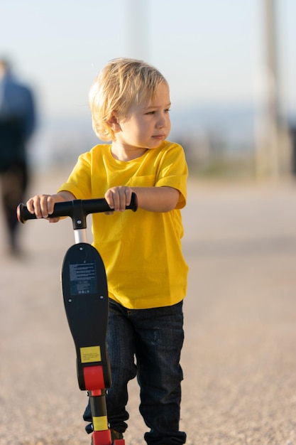 Un petit garçon se tient sur la route tenant par un scooter