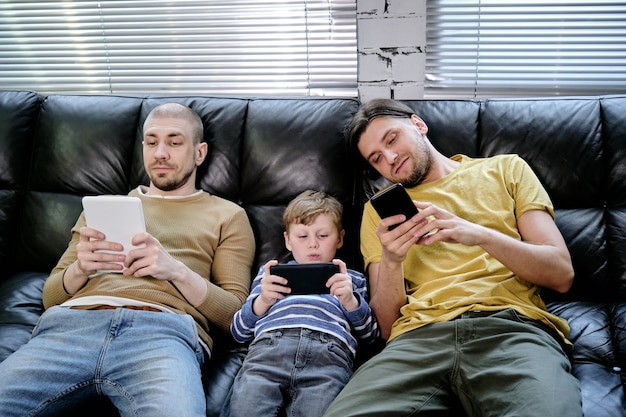 Petit garçon avec smartphone assis sur un canapé entre deux hommes avec des gadgets