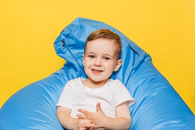 Petit garçon souriant assis sur une chaise bleue sur fond jaune vif