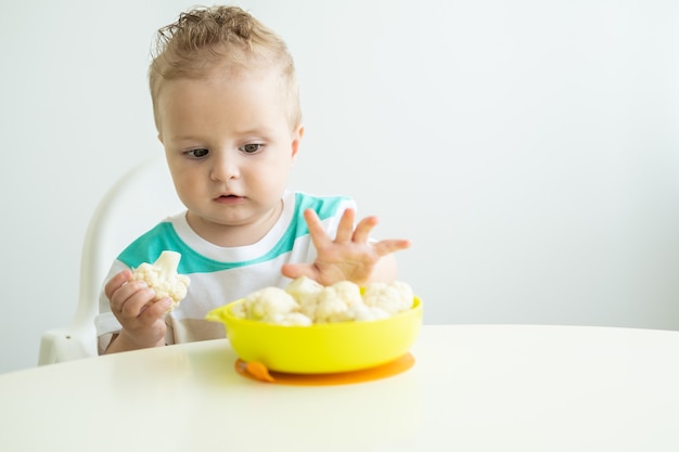 Petit garçon souriant assis dans une chaise d'enfant mangeant du chou-fleur sur une cuisine blanche.