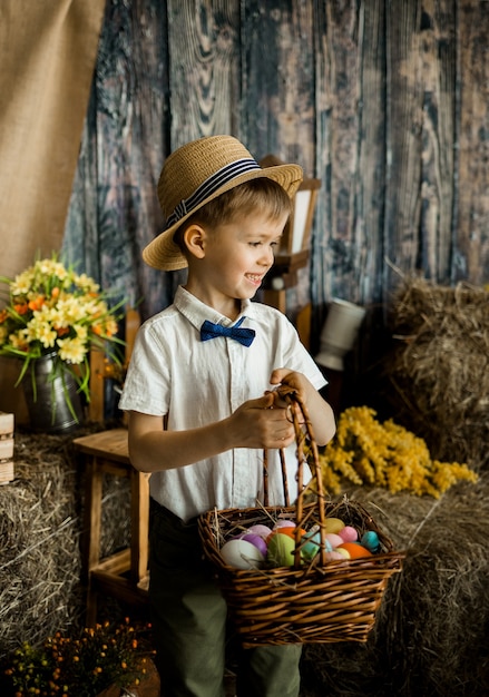 Petit garçon souriant dans une chemise, un noeud papillon et un chapeau de paille se dresse avec un panier d'oeufs colorés dans les décorations de Pâques