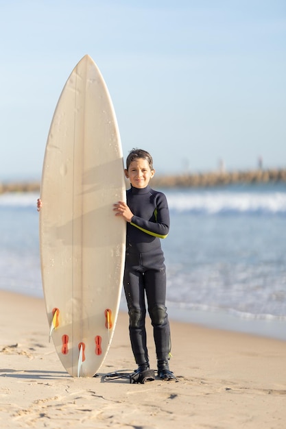 Photo un petit garçon souriant debout au bord de la mer avec une grande planche de surf