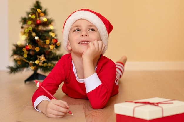 Un petit garçon souriant en vêtements de Noël écrit une lettre au Père Noël allongé sur le sol. Notion de Noël et des enfants.