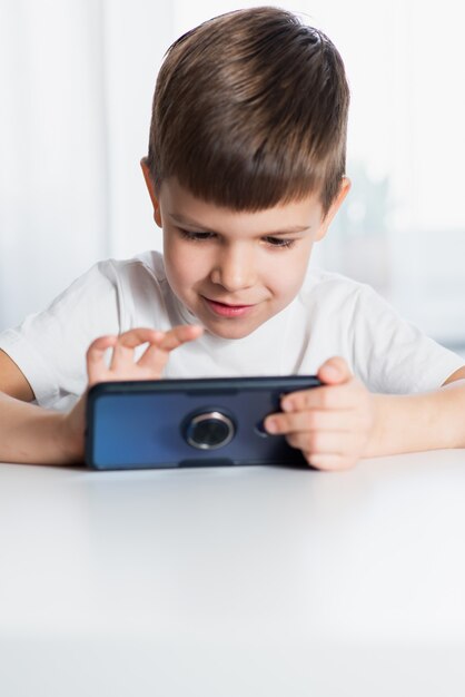 Un petit garçon en T-shirt blanc joue à des jeux au téléphone à la maison. Un enfant heureux regarde son smartphone.