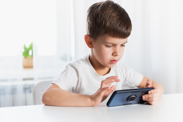 Un petit garçon en T-shirt blanc joue à des jeux au téléphone à la maison. Un enfant heureux regarde son smartphone.