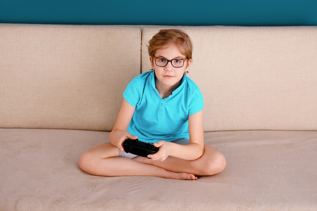 Un petit garçon en T-shirt bleu et de grandes lunettes assis sur le canapé et jouant à la maison avec une manette de jeu