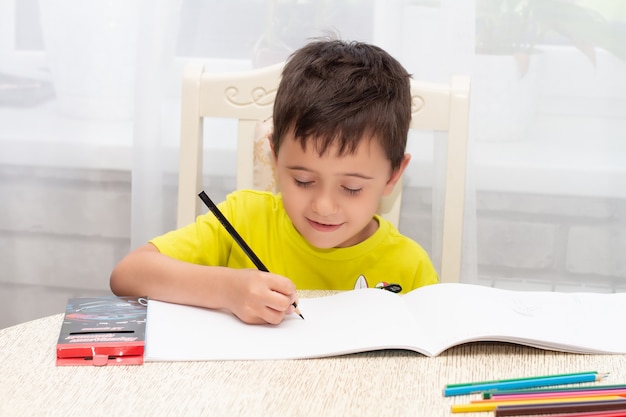 un petit garçon en T-shirt jaune est assis à la maison à la table et dessine avec des crayons un album