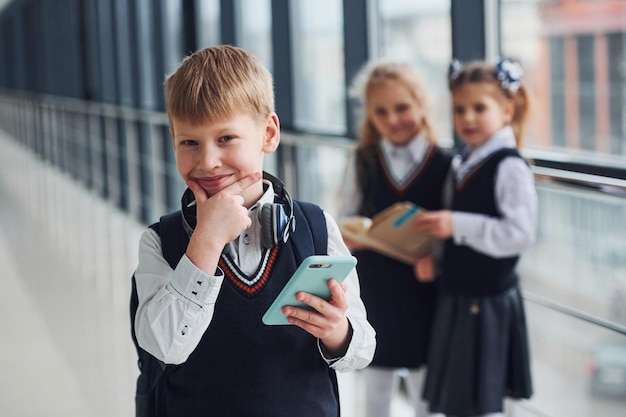Petit garçon avec téléphone et casque debout si devant des écoliers en uniforme qui ensemble dans le couloir Conception de l'éducation