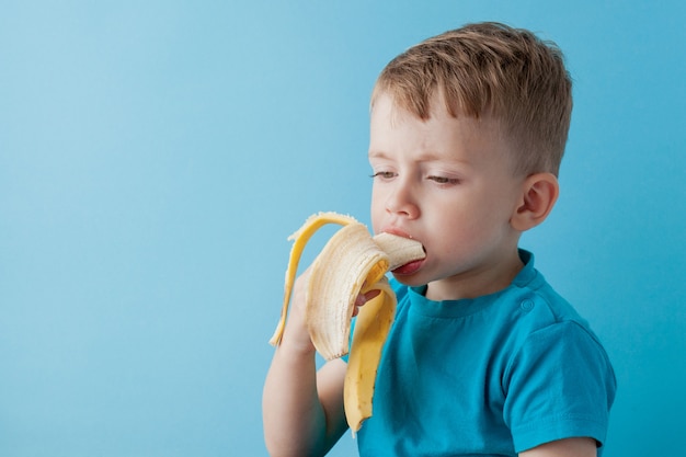 Petit garçon tenant et manger une banane sur le mur bleu, nourriture, alimentation et concept de saine alimentation