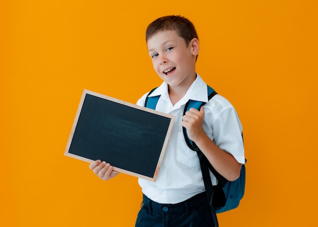 Petit garçon tenir le tableau noir de l'école sur fond jaune. Petit uniforme scolaire d'écolier avec un sac à dos avec les mains vides.