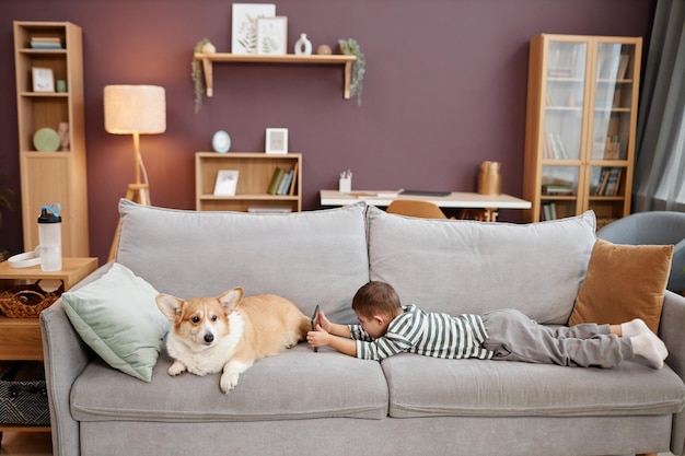 Petit garçon utilisant une tablette jouant à des jeux mobiles allongé sur un canapé avec un chien