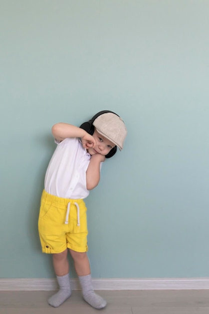 Un petit garçon vêtu d'une chemise blanche, d'un short jaune et d'une casquette rétro écoute de la musique à travers de gros écouteurs