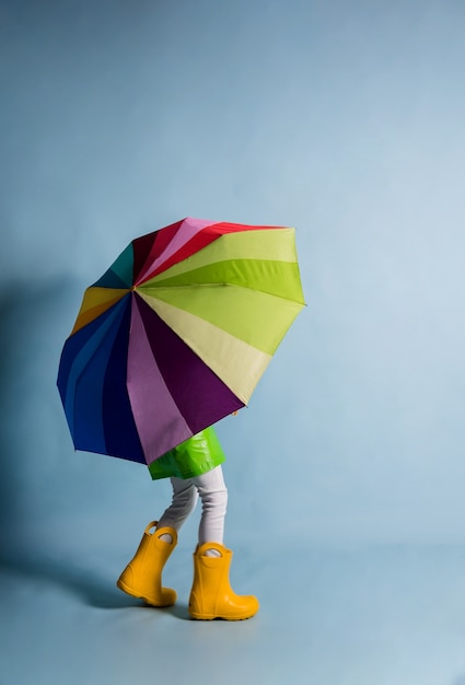 Un petit garçon vêtu d'un imperméable vert et de bottes en caoutchouc jaune s'est couvert d'un parapluie multicolore sur fond bleu avec une place pour le texte