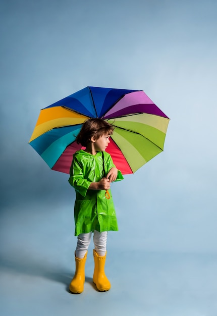 Un petit garçon vêtu d'un imperméable vert et de bottes en caoutchouc jaune tient un parapluie multicolore sur fond bleu avec une place pour le texte