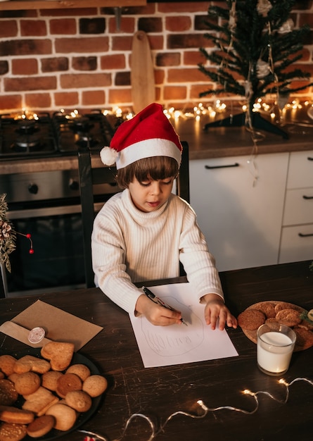 Un petit garçon vêtu d'un pull en tricot blanc et d'un chapeau rouge du Nouvel An est assis dans la cuisine et écrit un message du Nouvel An