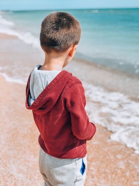 Un petit garçon vêtu d'une veste et d'un pantalon se tient au bord de la mer avec son dos et regarde vers l'eau. Concept de loisirs et de tourisme.