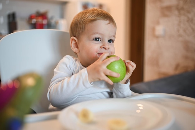 Petit heureux mignon bébé garçon blond assis sur une chaise de bébé jouant avec le visage de bébé pomme