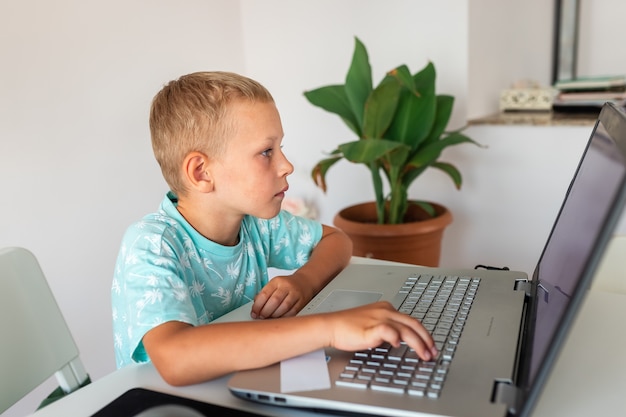 Petit jeune écolier travaillant à la maison avec un ordinateur portable et des notes de classe étudiant dans une classe virtuelle.