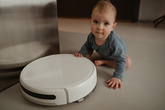 Petit et mignon bébé sur de la farine jouant avec un aspirateur robot à la maison.