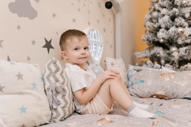 Petit mignon petit garçon assis dans la chambre des enfants dans une maison de lit en bois avec des veilleuses en forme de ballon à côté d'un arbre de Noël