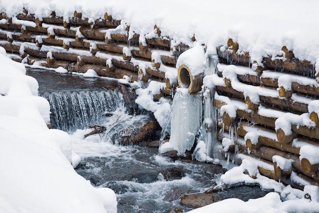 Un petit mur de petites bûches entoure un petit ruisseau de montagne avec des cascades dans une forêt d'hiver près d'arbres nus dans les montagnes des Carpates