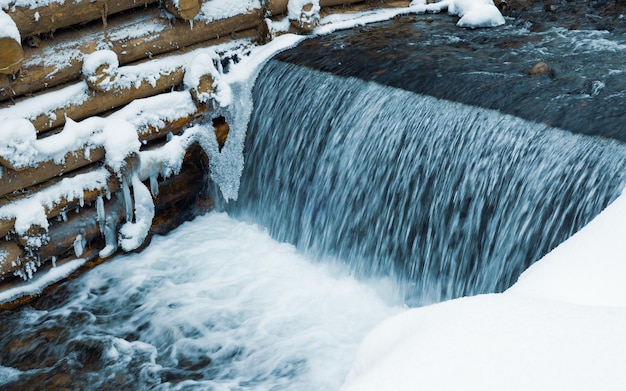 Un petit mur de petites bûches entoure un petit ruisseau de montagne avec des cascades dans une forêt d'hiver près d'arbres nus dans les montagnes des Carpates