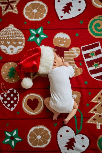 Un petit nouveau-né endormi, portant un bonnet de Noel et un body blanc vierge, se trouve sur une couverture festive de Noël rouge avec une guirlande. Vue de dessus, maison de Noël à l'intérieur