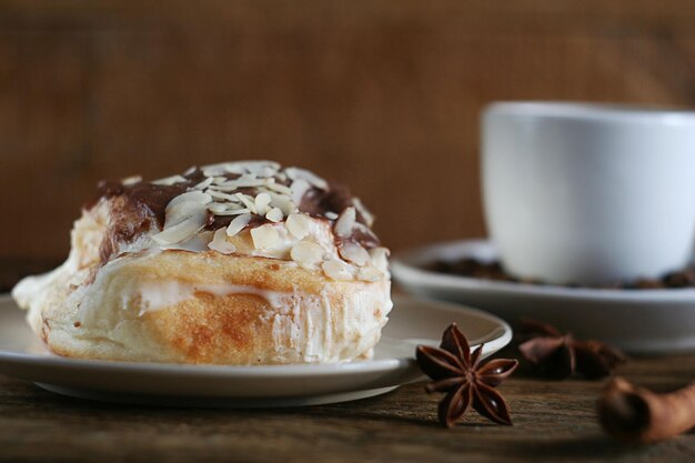 Photo un petit pain sucré avec du chocolat à la crème et des noix se trouve dans une assiette sur une table en bois derrière se trouve une tasse de café à proximité se trouvent des bâtons de cannelle et de l'anis étoilé