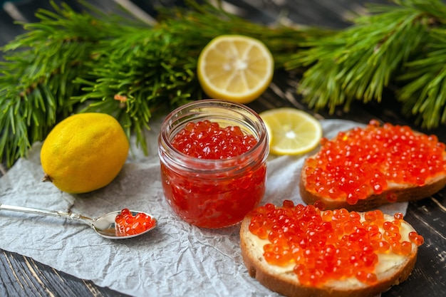 Petit pot en verre avec caviar rouge, citrons et aiguilles sur une table en bois noir