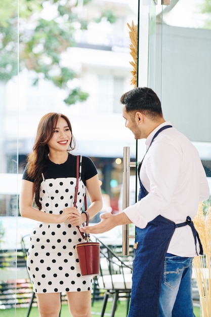 Petit propriétaire de boulangerie ouvrant la porte pour jolie femme, faisant un geste de bienvenue et l'invitant à l'intérieur