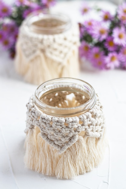 Petit vase en verre, pot, bougeoir avec couvercle en macramé. Fleurs d'automne floues sur fond. Bohème