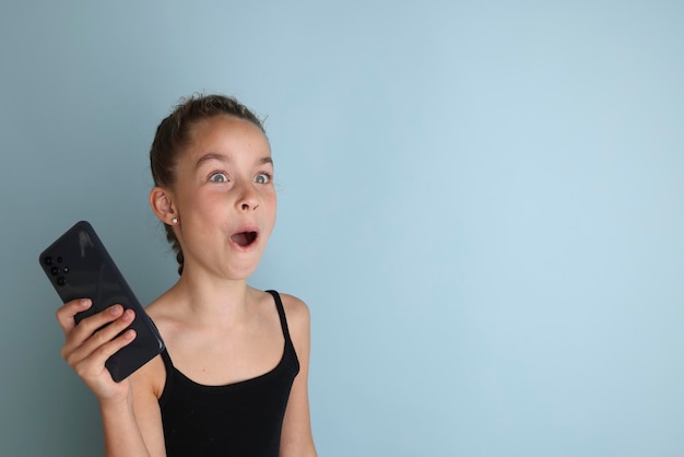 Petite adolescente émotive dans un t-shirt noir 11 12 ans sur un fond bleu isolé avec un smartphone dans ses mains Portrait en studio Children39s Placez le texte pour copier l'espace pour la légende